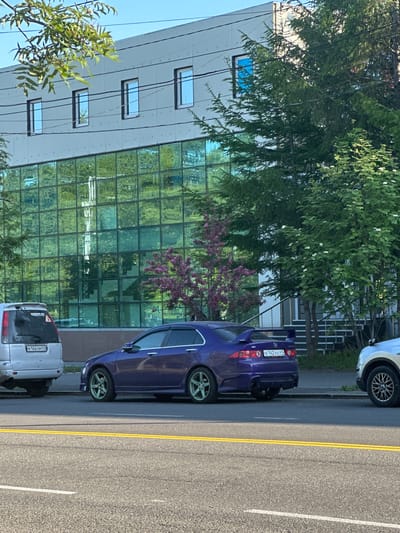 In Yelizovo, Russia, a purple sports car with a prominent rear spoiler is parked on the side of a street. The car has distinctive light green rims. To its left, a silver SUV is parked. In the background, a modern building with a green, reflective glass facade stands tall. Trees, one with pink blossoms, are interspersed around the building and along the street. Power lines are visible running across the facade of the building. The street is paved with asphalt and marked with yellow and white lines. The sky above is clear and blue.