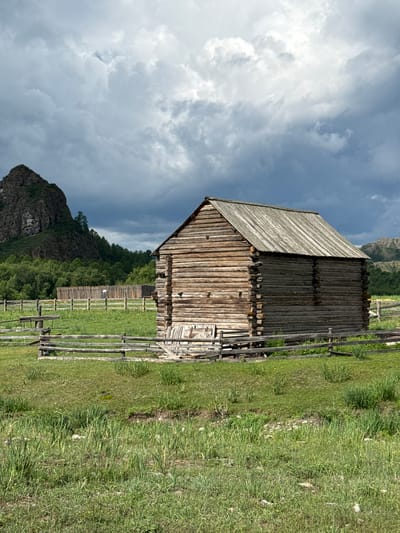 The image depicts a rustic wooden structure, likely a barn or storage building, situated in a rural landscape. The building is constructed from rough-hewn logs, with a weathered wooden roof. It is enclosed by a simple wooden fence that demarcates a grassy area. In the background, a large, rocky mountain peak rises against a dramatic, cloudy sky. Dense green foliage, including trees, forms a band between the fenced area and the mountain. Further in the distance, more rolling, forested hills are visible. The foreground consists of a field of tall, green grass. The overall scene suggests an outdoor, natural environment, possibly a farm or a remote settlement, in the area of Kazanovka, Russia. There are no people visible in the image.
