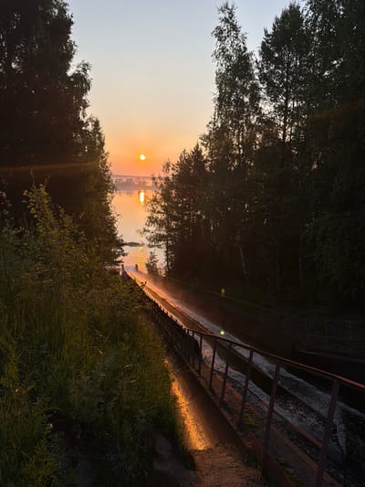 An early morning sunrise illuminates a concrete channel with a metal railing on the right. The channel appears to be a spillway or canal, with water flowing down it, creating a misty spray. The sun is low on the horizon, casting a warm golden light on the water and the surrounding trees and vegetation. Lush green grass and foliage are visible on the left side of the frame, illuminated by the sunrise. The sky is a clear gradient of pale blue to soft orange. The location is the city of Chernushka, Russia, at the water outlet from the Chernushka reservoir. No people are present in the image.