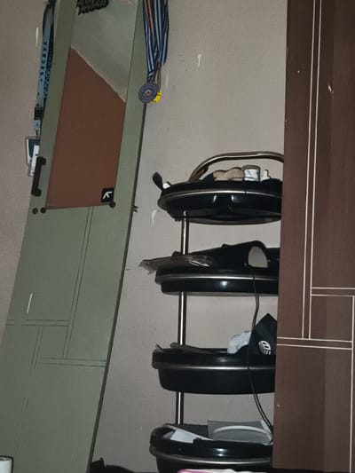 The image captures an indoor scene, likely a personal living space such as a room or hallway, in Pacet, Indonesia. No people are present.
On the left, a tall, light-green panel, possibly a door or a large mirror, leans against a light-colored wall. This panel has a distinct reddish-brown rectangular section and subtle grey line patterns. Hanging from its top left are a blue lanyard with white text, partially visible as "SUMMIT SENIOR", and a blue and white striped medal. The medal's circular medallion clearly shows "SATU" on its inner ring, with additional, unreadable text on the outer ring. A small black sticker with a white stylized 'K' or similar geometric symbol is affixed to the lower right of this green panel.
To the right, a three-tiered storage rack, likely a shoe rack, is visible. It features a metallic, possibly gold-toned, frame supporting black circular trays. These trays hold various items, predominantly footwear such as slippers, sandals, and sneakers, along with charging cables and other small personal belongings. On the second shelf from the bottom, a white item displays a black rectangular logo with white text resembling "DG" or "D II". The entire right side of the frame is partially obscured by a vertical brown wooden door or wall panel, characterized by two thin white vertical lines.
The lighting in the scene is dim, suggesting it was taken during the evening or in an interior space with limited ambient light. The arrangement of objects conveys a sense of everyday use and personal organization within the domestic setting.