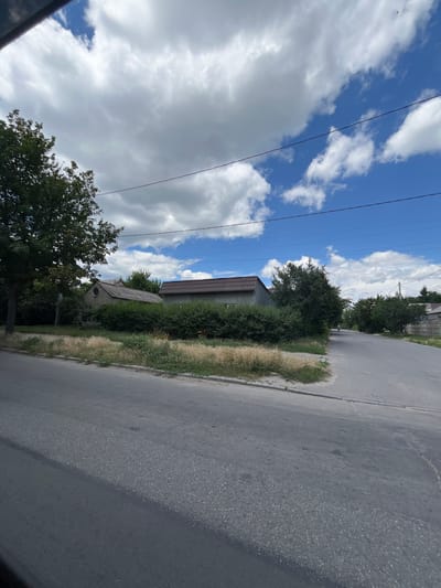 This is a vertical, low-angle outdoor shot of a street in Dnipro, Ukraine. The sky is a vibrant blue, dotted with fluffy white clouds and crisscrossed by power lines. To the left of the frame, a dense green tree grows tall against the sky. In the middle ground, a low-slung building with a brown tiled roof is partially visible behind a neatly trimmed hedge. Further back, another structure with a sloped roof and a round window stands amidst lush greenery. The right side of the image is dominated by a grey asphalt road, bordered by a strip of dry, light-brown grass and a thin concrete curb. The overall scene suggests a sunny day, possibly late morning or early afternoon, in a semi-rural or suburban setting. No people are visible in the frame.