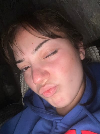 A person with light skin lies tilted on a dark surface wearing a blue hoodie. Their dark brown hair is visible along their forehead and around their face. One eye is closed, and the other is slightly open revealing a dark iris. Their lips are pursed. Part of a white woven object is seen near their head.