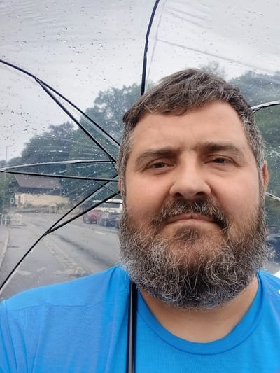 A man is taking a selfie under a clear umbrella. The man has a full beard with graying hair. He is wearing a bright blue t-shirt. The umbrella is clear and has raindrops on it, indicating it is raining. Through the umbrella, trees and buildings can be seen, suggesting an outdoor setting in a town or city. The man's expression is neutral. The lighting suggests it is daytime, likely overcast due to the rain. The context provided indicates the location is Pfarrkirchen, Germany.