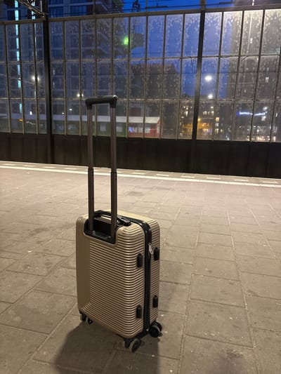 A gold, wheeled suitcase stands on a paved platform next to train tracks. The suitcase is positioned in the foreground, angled slightly towards the left, with its handle extended upwards. The platform is made of large, grey concrete pavers. Behind the suitcase, a dark, metal wall runs horizontally, partially obscured by the suitcase. Above the wall is a large, grid-like window structure, through which the lights of a city at dusk are visible. The windows are speckled with condensation or dirt, obscuring the view of the buildings behind. The sky above is a deep blue, suggesting twilight. The overall scene appears to be a train station, likely in an urban setting. Considering the specific mention of Eindhoven, Netherlands, this scene likely depicts a platform at Eindhoven Central Station. There are no people visible in the image.