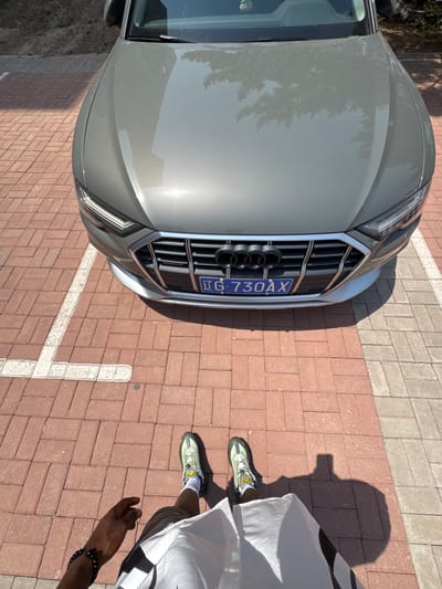 An overhead shot captures a gray Audi car parked on a paved surface of red and gray interlocking bricks, with a person standing beside it. The car, likely an SUV or crossover, faces towards the viewer with its distinctive four-ring Audi emblem prominent on the front grille. A blue Chinese license plate, bearing the characters "京 G.730AX," is mounted below the grille, indicating the car is registered in Beijing. Reflections of trees are visible on the car's shiny hood, suggesting an outdoor setting.

To the right of the car, a person's lower body is visible, wearing light-colored shorts and a white garment with black patterns. They stand on the brick pavement, which features a white painted parking line. The person is wearing light green/gray athletic shoes with yellow accents and a small yellow emblem on the side. A dark-skinned hand and forearm, adorned with a black beaded bracelet, are also visible towards the bottom right of the frame.

Bright sunlight creates sharp shadows of the person and the car on the ground, indicating a clear, sunny day, likely mid-morning to afternoon. The scene takes place in an outdoor parking area in Zhongtun, China, as suggested by the license plate and provided context. No specific activity is depicted other than standing next to the vehicle.