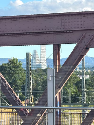 This image captures a view from a modern bridge, likely the Dreiländerbrücke in Weil am Rhein, Germany, looking across a valley on a clear, sunny day. The dominant subjects are the reddish-brown metal girders of the bridge, characterized by numerous rivets, suggesting robust construction. These structural elements frame the composition, with a large beam running along the upper part and diagonal supports extending into the view.

Through a transparent or reflective barrier (possibly glass), the scene beyond the bridge is visible. To the upper right, a bright blue sky with scattered white clouds dominates. Below this, a verdant landscape unfolds, featuring dense green trees and distant hills or mountains. In the middle ground, several tall, modern buildings with repetitive window patterns stand out against the horizon, indicating an urban area, likely Basel, Switzerland. Another, lighter green metal bridge structure, possibly a railway bridge or part of the overhead railway system, is also visible. A subtle grid pattern, likely a reflection from a mesh or fence on the viewing platform, overlays parts of the scenery. A silver vertical pole, possibly a handrail, is present on the right side.

No specific activity is taking place within the frame, but the image suggests the experience of observing the landscape from the bridge. The weather is bright and sunny.

Visible text on a distant billboard through the reflection is partially discernible as "SPARKAS", strongly suggesting "Sparkasse," a German banking group.