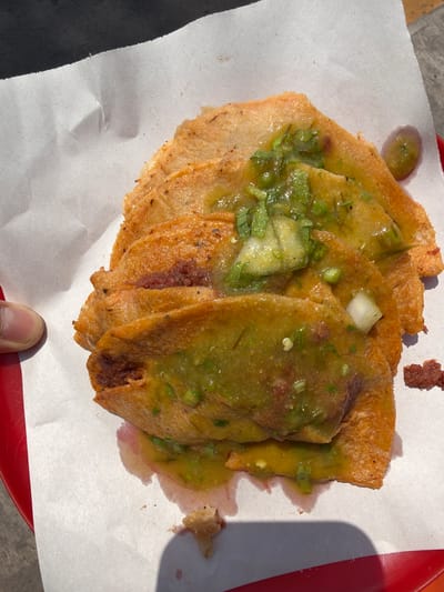 The image shows a plate of golden-brown tacos dorados (crispy tacos) covered in green salsa. The tacos are arranged on a white paper liner, which rests on a red tray. A fingertip is visible at the bottom of the frame, near the tacos. The photograph was likely taken in Puebla, Mexico, as tacos dorados are a regional specialty. The tacos appear to be freshly prepared and served.
