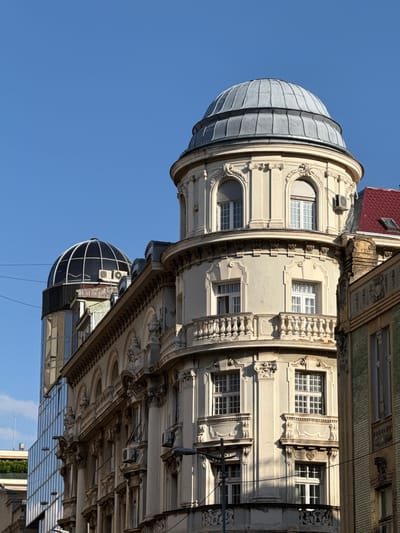 The image shows the facade of the Hotel Moskva in Belgrade, Serbia. The hotel is a landmark building located at the intersection of Terazije and Balkanska streets. The image focuses on the upper floors of the hotel, particularly a distinctive curved corner with a dome. The building exhibits architectural details typical of Art Nouveau and Secessionist styles, with ornate decorations, arched windows, and a curved balcony. A modern glass building is visible to the left of the Hotel Moskva, creating a contrast between old and new architecture. The sky is clear and blue, indicating a sunny day. No people are visible in the image.