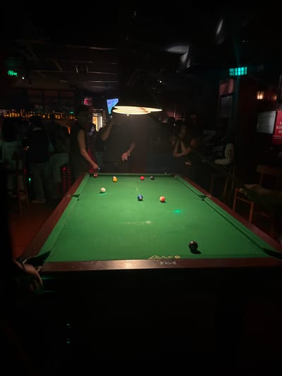 A dimly lit interior scene shows a pool table with several people around it. The location appears to be a bar or recreational establishment. The pool table is green, and several billiard balls are scattered across its surface. People are standing around the pool table. The lighting is poor with several sources of lighting. The location may be in Liangjiang, China, though no specific clues from the environment are visible.