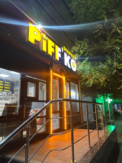 The image shows the exterior of a market named "Piffko" at night. The market has a bright yellow illuminated sign. A metal railing leads up to the entrance, which is lit by vertical orange lights. The street and sidewalk are visible, along with trees and other buildings. The scene appears to be in the city of Naberezhnye Chelny, Russia.