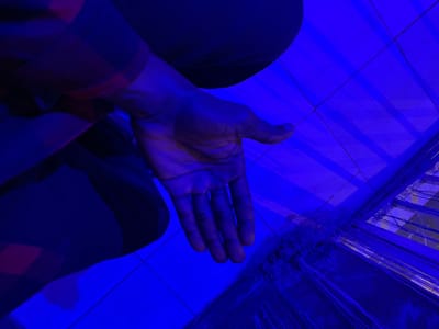 In Durumi, Nigeria, an individual is positioned with their left leg bent and their right hand extended forward, palm open. The person is wearing a checkered shirt and dark trousers, bathed in a vibrant blue light that casts strong shadows. The background consists of a tiled floor with a visible grate in the lower right quadrant of the image. The overall lighting creates a dramatic and somewhat abstract visual.