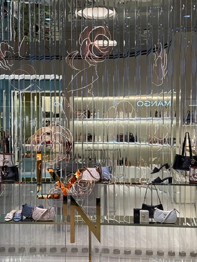 The image depicts a retail store interior, likely a clothing or accessories boutique, with a focus on handbags displayed on shelves behind a reflective, horizontal-slatted partition. The store appears to be modern and well-lit, suggesting it's open for business.

The primary objects of interest are various handbags in different styles, colors, and sizes. Some are displayed prominently, while others are partially obscured by the reflective slats or other items. The reflective surface creates a layered and somewhat distorted view of the merchandise, with multiple reflections and a sense of depth.

There are also decorative elements in the store, including illuminated line art of flowers and the brand name "MANGO" rendered in neon or light tubing, which is reflected in the partitioning. A large, abstract golden "Z" shape is also visible, adding to the store's aesthetic. The lighting suggests it could be daytime or evening, as the interior is brightly lit, and there are no external weather cues.

The overall scene conveys a sense of commercial display and visual merchandising, aiming to attract customers. The reflections add an element of artistic interest to the otherwise straightforward presentation of goods. The location context of Wichit, Thailand, is a factual detail about the setting. There are no visible people in the image.