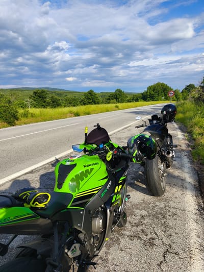 Two motorcycles are parked on the shoulder of an asphalt road in a rural area. The foreground motorcycle is a green and black Kawasaki ZX-10R with gloves resting on the seat. A black helmet is on the handlebars of the motorcycle. The background motorcycle is black with a black helmet on the handlebars. The road extends into the distance with green fields and trees on either side. In the background, there are rolling hills. A red and white 'no passing' sign is posted near the background motorcycle. The sky is blue with puffy white clouds. The location is Mladinovo, Bulgaria.