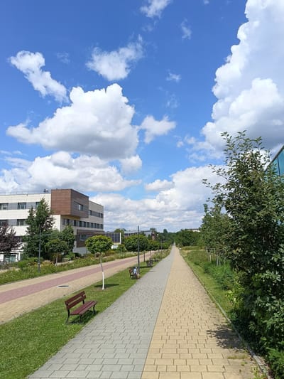 The image depicts a sunny day with scattered clouds over a modern building complex. A paved walkway stretches into the distance, flanked by green grass and trees. Benches are placed along the walkway, with one person seated on a bench in the distance. The architecture suggests an institutional or corporate setting. This scene is characteristic of the area around the Jagiellonian University's campus in Krakow, Poland, specifically the northern part of the city, known for its modern research and educational facilities.