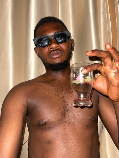 A dark-skinned man wearing sunglasses lies on a satin sheet. His head is tilted back. A hand holds a small, clear vase filled with flowers over his bare chest. The vase's scalloped edge is visible. The man has a dark beard and short hair. His lips are slightly parted. The lighting is soft and highlights the textures of the skin and fabric.