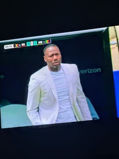 The media file shows a television screen displaying a soccer match. A man in a white suit is visible on the screen, looking concerned. The screen displays a scoreboard with the time "09:18" and the score "0-0" between "BEL" (Belgium) and "SEN" (Senegal). There is also a "Verizon" logo visible on the screen. The image appears to have been taken indoors in Gwarinpa, Nigeria, given the location context provided. The overall mood seems to be tense, possibly due to the close score and the man's expression.