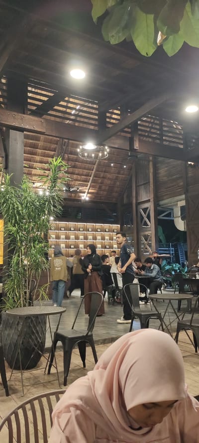 This media file depicts an indoor gathering at a cafe with a rustic, wooden-themed interior. The lighting suggests it is evening or night. Several people are present, engaged in various activities. In the foreground, a person wearing a light pink hijab is seated, partially visible.
Further back, a group of young adults are interacting. One man, wearing a dark t-shirt with a graphic design and dark pants, stands looking to the side. Next to him, a woman in a black hijab and a long, brown skirt appears to be speaking with him or someone off-camera. Other individuals are seated at tables, some appearing to be in conversation.
The cafe's architecture features exposed wooden beams and a high, sloped ceiling with visible rafters, creating a spacious and somewhat industrial feel. A large chandelier hangs from the ceiling, and several circular lights are also visible, illuminating the space. Lush green plants are arranged near a round, dark metal table and chairs in the left foreground, adding a touch of nature to the setting. In the background, shelves lined with small, square items, possibly books or framed pictures, are mounted on the wall. The overall atmosphere seems casual and social. There is no visible text in the image.