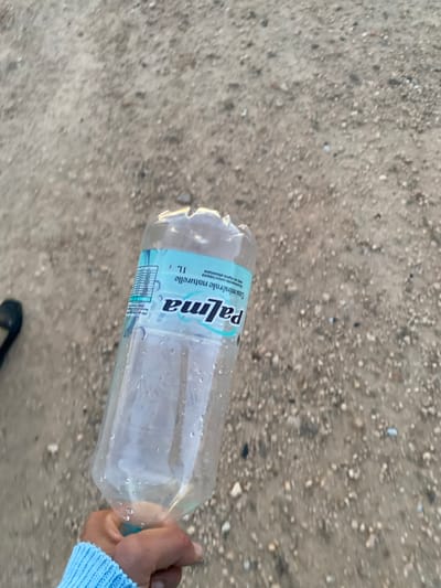 A clear plastic water bottle, labeled "Palma" and "1L", lies horizontally on a textured gray and brown ground. Condensation droplets are visible on the bottle's surface. A hand wearing a light blue sweater holds the bottle's end. A black shoe is partially visible in the lower center of the frame. The background appears to be a dirt or gravel surface with varying shades of gray and brown.