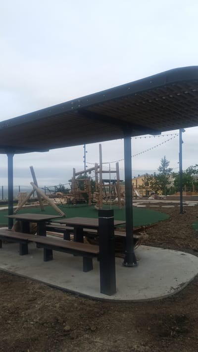 An outdoor view of a playground, featuring a covered seating area with picnic tables and benches, is shown. The playground equipment, made of wood and rope, is visible in the background. The scene is set against a backdrop of a cloudy sky and a distant cityscape with buildings and trees. The ground is a mix of dirt and green turf, indicating ongoing landscaping or construction. The location is identified as Vladivostok, Russia.