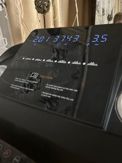 The scene depicts a close-up, slightly elevated view of a black, glossy treadmill control panel located in what appears to be an indoor residential setting in Dmitrievskaya, Russia. The panel displays blue LED readouts: "2:01", "3743", and "3.5", which are labeled with "TIME" and "SPEED" icons, indicating an elapsed time of 2 minutes and 1 second, a metric value of 3743 (e.g., distance or calories), and a speed of 3.5 (e.g., mph or km/h). Below these figures, a series of horizontal bar graphs suggests program profiles or intensity levels. Further down, a "CAUTION" section, accompanied by an open book icon, lists four safety instructions: "Read the owner's manual carefully before using the product," "Keep children away from the treadmill during operation," "To avoid injury, stand on the side rails before starting the unit," and "Remove the safety key when the unit is not in use." The glossy surface of the control panel reflects elements from the background, including light-colored curtains, a dark, spherical decorative object on a slender stand, and the faint upper part of a person's head with light-colored hair, visible in the upper right quadrant of the reflection. The treadmill appears to be in an operational or recently used state.
