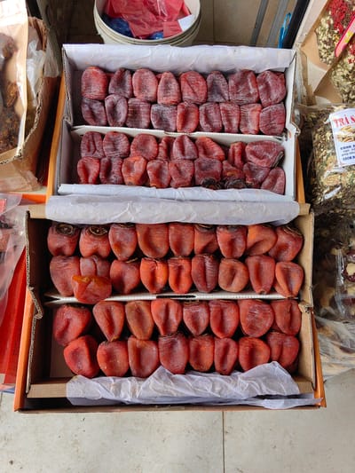 The image displays an overhead view of a market stall in Sa Pa, Vietnam, showcasing various dried food products. The central focus is a two-tiered arrangement of dried persimmons within stacked corrugated cardboard boxes. The top box contains two layers of shriveled, reddish-brown dried persimmons, some with a faint white powdery coating, arranged in neat rows. The bottom box similarly holds two layers of the same dried fruit. White crumpled tissue paper separates some of the persimmon layers within the boxes.
To the immediate left of the boxes, a brown paper bag contains a translucent plastic bag filled with dark, irregularly shaped dried items. Further left and above, a white plastic bucket holds red and blue plastic bags. To the right of the persimmon display, multiple clear plastic bags are visible, containing various dried herbs and floral components. One prominent bag features a white label with red and black Vietnamese text, partially visible as "TRÀ S..." (likely indicating "Sapa Tea" or similar). Other bags contain what appear to be dried jasmine flowers, red flower petals, and reddish-brown dried fruits or berries. The entire display rests on a light-colored, possibly tiled or concrete, ground surface. No individuals or specific actions are depicted.