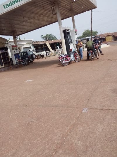 The scene depicts a daylight view of a gas station located in Ankpa, Nigeria. A large, flat canopy, partially displaying "Yadali" in green text, extends over a reddish-brown asphalt or concrete fueling area. Under the canopy, multiple fuel pumps are visible; one pump clearly displays a "NO SMOKING" warning.
Four individuals are present. One person, wearing a patterned headscarf and blue jeans, stands near a fuel pump, appearing to operate it or attend to a red and white motorcycle parked beside it. To their right, a man in a green shirt is seated on a dark motorcycle, positioned near another pump. Behind him, a second man in a dark blue shirt is seated on another dark motorcycle. A fourth person is partially obscured by the man in the green shirt.
Vehicles include the red and white motorcycle, two dark motorcycles with riders, a dark SUV or pickup truck parked near a white building under the canopy, and a small white utility truck with an attached trailer in the background. Behind the main fueling area, a white building stands under the canopy, with several older structures featuring corrugated iron roofs visible further back. Green foliage is present in the distant background under a hazy sky.