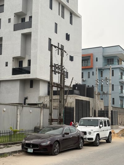 The image captures an urban street scene in what appears to be a residential or commercial area. On the left, a prominent modern building with a textured white facade stands tall, featuring numerous square and rectangular windows, some recessed. Behind it and to the right, another multi-story building with a light blue-grey exterior is visible, also exhibiting a modern architectural style with multiple windows and visible air conditioning units.
In the mid-ground, a dense network of utility poles and electrical wires stretches across the frame, indicating an active power infrastructure. Several black, cylindrical tanks (likely water storage) are situated on the ground level near these utilities, with one tank displaying a symbol resembling a stylized 'H'.
Parked on the right side of the street are two luxury vehicles. In the foreground, a dark-colored (possibly maroon or dark brown) Maserati sedan is visible, its front grille and headlights clearly in view. The license plate on this car reads "AGL 148GY" and "LAGOS," confirming the location as Lagos, Nigeria. Behind the Maserati, a white Mercedes-Benz G-Wagen SUV is parked. A person wearing a red shirt is partially visible behind the Maserati, near the G-Wagen.
The day appears to be overcast or cloudy, as indicated by the diffused lighting and lack of sharp shadows. A paved road occupies the right side of the image, while a short metal fence and some sparse green plants are seen along the roadside. The overall scene depicts a blend of modern architecture and infrastructure common in urban Nigerian settings.