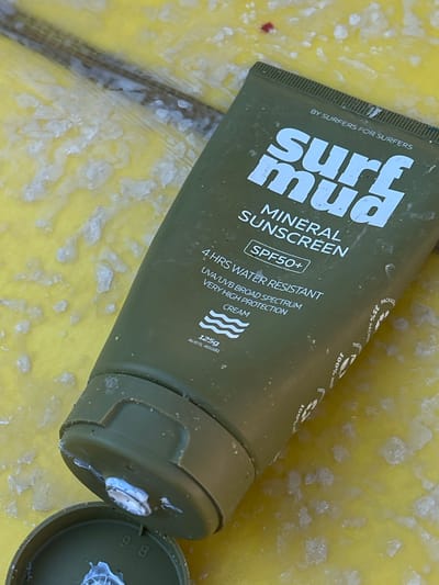 A close-up, eye-level shot shows a green tube of "surf mud MINERAL SUNSCREEN" lying on a yellow, textured surface with white specks. The tube is partially open, with a white cap lying beside it. The sunscreen label is visible, including the brand name, "MINERAL SUNSCREEN," "SPF50+", "4 HRS WATER RESISTANT," and "VERY HIGH PROTECTION CREAM." There are also indications of Australian origin, with "125g AUSTR 400381" printed on the tube. The background appears to be a surfboard or a similar surface, suggesting an outdoor environment.