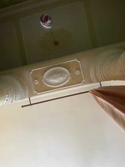 This media file shows an indoor scene looking upwards towards a ceiling adorned with decorative architectural elements. The primary subjects are the ceiling and its features, including ornate molding and a mosaic glass lampshade. The scene depicts a static view of the interior, with no people or specific activities evident.
Notable details include the detailed plasterwork on the ceiling, featuring intricate patterns and layered moldings. A unique lampshade, constructed from small, multi-colored mosaic tiles, hangs from a thin rod. A portion of a draped fabric, possibly a curtain or canopy, is visible in the upper right of the frame. The lighting suggests it is daytime, with soft light illuminating the ceiling. The location is identified as Ternopil, Ukraine, though no specific environmental cues are visible to confirm this beyond the provided context. There is no visible text within the image.