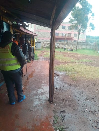 The image shows a scene in Gitaru, Kenya, located at the coordinates -1.218 latitude and 36.655 longitude. Three men are standing under a covered porch. The man in the center is wearing a blue shirt and a black jacket. The man on the left is wearing a yellow safety vest and black jeans. He is the subject of the image. He is standing closest to the camera. The man on the right is wearing a light-colored shirt and a dark jacket. He is standing to the right of the other two men and is partially obscured by the man in the center. The background of the image is a dirt lot with a large building in the distance. The building appears to be made of red brick and has a fence in front of it. The porch floor is made of concrete. The area is wet, likely from rain. The image appears to have been taken at a bus stop or bus station.