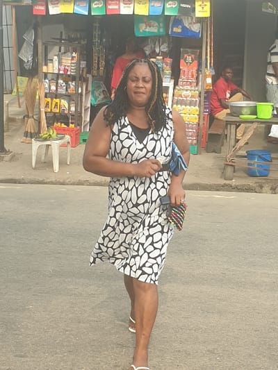This image captures a daytime street scene in Umuode, Nigeria, featuring a woman walking on an asphalt road in the foreground, with a roadside shop or stall in the background.
The primary subject is a woman of medium complexion, walking towards the viewer. She has dark hair styled in braids with visible lighter streaks and wears a sleeveless dress with a distinctive black and white abstract pattern over a black undershirt. Her footwear consists of white flip-flops. Her expression appears solemn or serious. In her left hand, she holds a crumpled light blue cloth. In her right hand, she carries a small, multicolored beaded clutch purse alongside a dark rectangular object, possibly a mobile phone.
The background consists of a storefront. Above the entrance, various product advertisements for brands such as Milo, Nescafe, Quality, Nido, Johnny, Indomie, and Maggi are displayed. The shop entrance reveals assorted merchandise, including shelves stocked with what appear to be motor oils or lubricants in yellow bottles, bundles of brooms, and stacks of packaged beverages in crates. A small, white plastic table to the left holds a bunch of green bananas. Inside the shop, a person in a red shirt is partially visible. To the right, a man in a red shirt is seated on a low wooden bench next to a larger wooden table that holds a large metal bowl and a green plastic bucket. A third individual, wearing a striped shirt, is partially visible standing on the far right. The overall environment suggests a commercial area along a main thoroughfare.