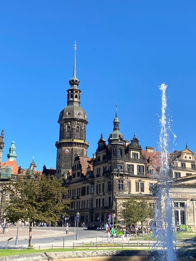 The image captures a view of Dresden, Germany, featuring the Dresdner Residenzschloss (Dresden Castle) with its Hausmannsturm tower rising prominently against a clear blue sky. A fountain in the foreground appears to spray water upwards, partially obscuring the view of the buildings directly behind it. The buildings are constructed with intricate architectural details and varying heights, suggesting a mix of historical styles.
The streets and sidewalks surrounding the castle show pedestrian activity, with people moving along the walkways. Trees are interspersed throughout the scene, adding greenery to the urban landscape. Lampposts line the street. The overall environment suggests a sunny day in a bustling city center, with the castle acting as a focal point and a landmark.