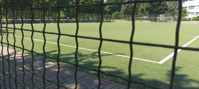 The image shows an outdoor artificial turf soccer field. The field has white markings indicating the boundaries and center circle. A soccer goal is visible in the distance. Bleachers are set up along one side of the field, with a fence separating the field from a paved walkway. Trees and a building are visible in the background. The photograph is taken from behind a metal mesh fence, which partially obstructs the view of the field. The scene appears to be in a park or sports facility within the city of Cherkasy, Ukraine.