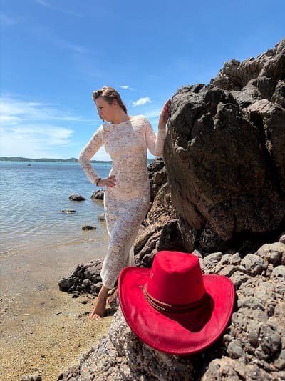 In Baan Bo Phut, Thailand, a woman in a white lace dress stands on a rocky beach with the ocean to her left and a large rock formation to her right. She is looking down and to her left, with her left hand on her hip and her right hand resting on the large rock. In the foreground, a red cowboy hat rests on the rocks. The sky is blue with scattered clouds, and the water is calm.