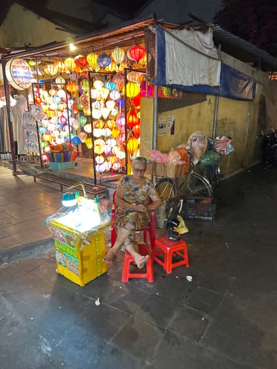 The image depicts a street scene at night in Hoi An Ward, Vietnam, characterized by a vibrant display of colorful lanterns. In the foreground, an elderly woman with glasses sits on a red plastic stool, her legs crossed. She is wearing a floral-patterned dress. To her left is a yellow, illuminated cart displaying food items and text in Vietnamese, including "BANH XOA" and "MANGO CAKE". A smaller red plastic stool is positioned to her right. Behind the cart, a bicycle is laden with baskets of goods and wrapped items. The background features a stall with numerous hanging lanterns of various colors and shapes, illuminated and creating a festive atmosphere. The stall also displays clothing on mannequins. The street is paved with dark tiles. The overall impression is that of a lively night market or street vendor scene.