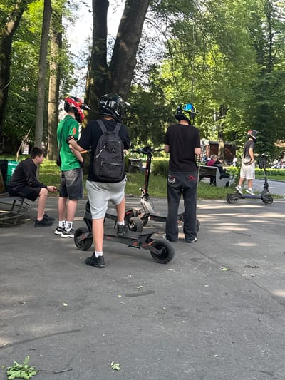 In Vinnytsia, Ukraine, a group of young people are gathered in a park, some wearing protective helmets and standing near electric scooters. The scene unfolds on a paved pathway, bordered by lush green trees and dappled sunlight. In the foreground, a young person in a black hoodie and shorts sits on a bench, holding a phone. Nearby, three other individuals stand, two of them wearing helmets and the third in a black t-shirt and dark jeans. They are positioned next to several electric scooters, suggesting an outing or a pause in their activities. Further back, other park-goers can be seen relaxing on benches amidst the trees. The atmosphere appears casual and recreational, typical of a sunny day in a city park.