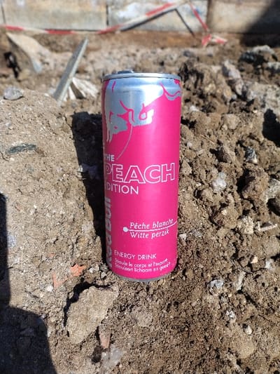 A pink and silver Red Bull "The Peach Edition" energy drink can, featuring text in French ("Pêche blanche") and Dutch ("Witte perzik"), stands upright on a pile of loose, dry brown soil mixed with small stones and debris. The can's surface shows a prominent silver bull logo. Bright sunlight illuminates the scene, casting a shadow to the lower left of the can. In the blurred background, piles of similar earth and fragmented construction materials are visible, alongside what appears to be a concrete structure and a red and white striped barrier tape. The scene is located in Crauthem, Luxembourg.