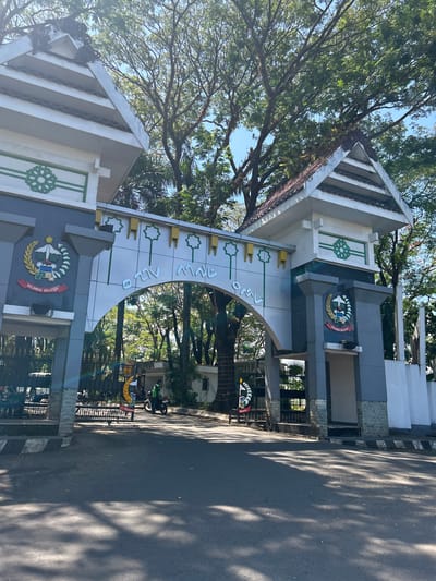 An arched gateway, featuring a government emblem and text in what appears to be a local script, marks the entrance to a facility in Makassar, Indonesia. The gateway is flanked by two structures with triangular roofs, adorned with decorative green patterns. Sunlight filters through the dense foliage of mature trees overhead, casting dappled shadows on the asphalt road in the foreground. A few motorcycles and people are visible beyond the gate, suggesting an active environment. The emblem on the left side of the gateway clearly reads "SULAWESI SELATAN", indicating the location of South Sulawesi.