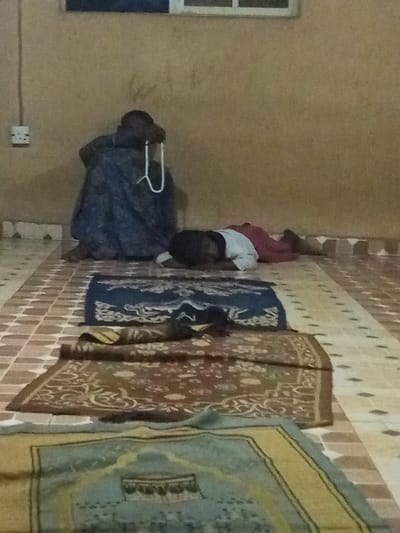 Inside a room, a person sits against a wall with a string of beads in hand. A child lies prone on a prayer rug in front of the person. Additional prayer rugs line the tiled floor. An electrical outlet is visible on the wall above the seated individual. A window is above the outlet. The wall appears to be a plain light brown color.