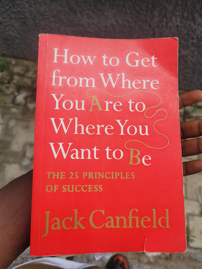 A person's hand holds a red book titled "How to Get from Where You Are to Where You Want to Be" by Jack Canfield, featuring "The 25 Principles of Success." The book's cover is bright red with gold lettering. The person's skin tone appears to be dark. The background is out of focus and shows a paved outdoor surface, possibly a street or walkway, with hints of vegetation or gravel. The overall scene suggests an outdoor setting in Katcha, Nigeria.
