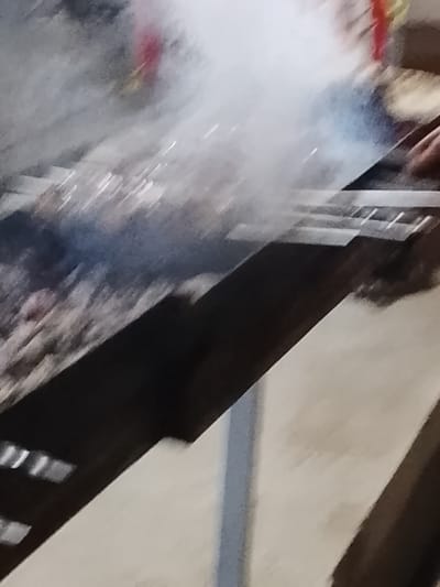 The image depicts a grill with meat cooking, producing a lot of smoke. The grill appears to be outdoors on a concrete or paved surface. The specific location within the city of Syrdarya, Uzbekistan, cannot be determined from the image.