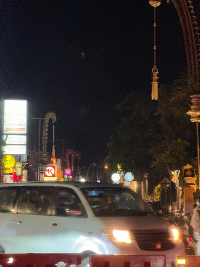This is a nighttime, medium-shot photograph taken in Ubud District, Indonesia. The scene depicts a street at night, characterized by illuminated signs, trees, and a moving vehicle. In the foreground, a white van is driving on the road, its headlights on. The van is partially obscured by a red barrier in the very bottom of the frame.
Behind the van, a variety of illuminated signs are visible. One prominent sign reads "Queen's of India Indian Cuisine" with a phone number. Other signs advertise "North & South Indian Cuisine, Veg & Non Veg" and "Pure 100% Indian Vegetarian". There are also signs for "Ubud" and a circular sign with a yellow crescent moon and stars. Further to the right, a red and white circular sign with a "K" is visible.
The background is dominated by dark trees and some decorative architectural elements, possibly part of a temple or archway. A tall, ornate hanging decoration, possibly a lantern or finial, is suspended from above. The overall atmosphere is that of a busy street at night, with commercial activity indicated by the numerous illuminated signs. The image is slightly grainy, consistent with low-light photography.