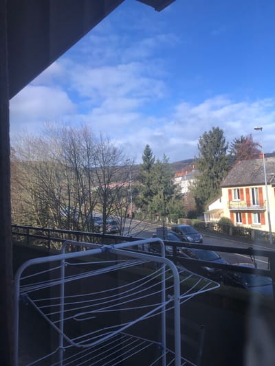 The image depicts an elevated outdoor view of a residential street in Saverne, France, under a bright blue sky with scattered white cumulus clouds. The perspective is framed by a dark overhead structure in the upper left and a dark metal balcony railing extending across the mid-foreground. A white, empty, multi-tiered clothes drying rack is positioned centrally in the lower foreground.

Below the railing, a paved street, appearing to curve slightly, runs from the left foreground towards the right. Several passenger vehicles are parked along the street, including a dark hatchback visible on the right side. On the immediate right, a two-story residential building with a light yellow facade, prominent orange-red window shutters, and a dark roof is visible. A grey street light pole stands to the right of this building. Further down the street, additional residential structures with lighter facades are observed.

The left mid-ground features a dense cluster of deciduous trees with bare branches, suggesting a winter or early spring season. Evergreen coniferous trees are interspersed among the buildings and on the distant landscape. The background consists of tree-covered hills or mountains, predominantly showing bare deciduous foliage. No individuals are present, and no actions or interactions are occurring within the scene. The environment is illuminated by direct sunlight, casting visible shadows.