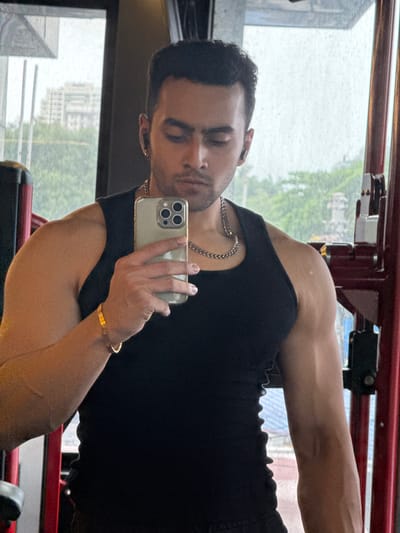 The image displays a man taking a mirror selfie inside what appears to be a gym or fitness center in Kolkata, India. The man has short, dark hair and light stubble, and is wearing a black sleeveless top. A silver-colored chain is visible around his neck. He holds a light-colored smartphone with a triple-lens camera setup in his left hand, angling it to capture his reflection. On his right wrist, he wears a gold-colored bracelet that appears to be inscribed with the word "LOVE" repeated.

The reflection is in a large window, offering a view of the exterior. Through the window, an urban landscape with multiple-story buildings and green trees or foliage can be seen. The window pane shows numerous small droplets, suggesting rainy or damp weather outside. Inside, behind the man, parts of red metal structures, consistent with gym equipment, are visible. The overall lighting indicates it is daytime. The man's expression is neutral to slightly serious.