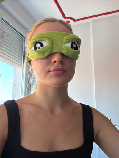 A young woman with blonde hair is photographed from the chest up, wearing a green eye mask with cartoonish eyes and a black tank top. She is indoors, with a window to her left showing blinds and a view of the sky. The background is a white wall with a red decorative element on the ceiling. The image is likely taken in a domestic setting within the city of Podgorica, Montenegro.