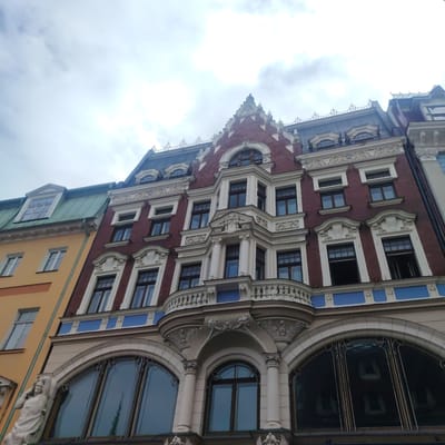 This media file captures an upward view of an ornate building facade in Riga, Latvia. The building displays a distinctive architectural style, featuring a red brick upper section with white decorative trim and a beige lower section. The facade is adorned with multiple windows, some with arched tops and elaborate detailing, and a prominent balcony with a decorative railing. On the left side of the image, a statue of a partially draped figure can be seen attached to the building's exterior. The sky above is cloudy, suggesting an overcast day. The image is a still shot and does not depict any ongoing activity or event.