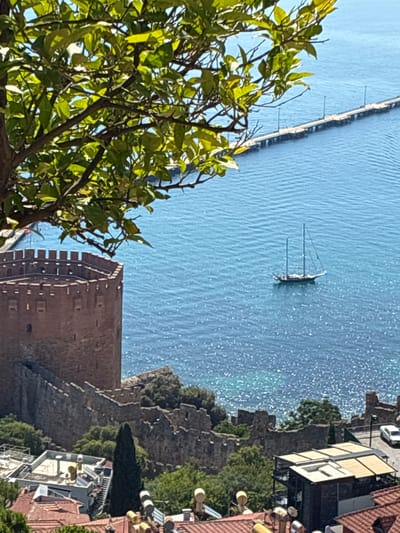 The image captures a scenic view of Alanya, Turkey, with a focus on its historic Red Tower and the surrounding coastline. In the foreground, the top branches of a leafy green tree partially obscure the view. Below the tree, the octagonal, red-brick Red Tower stands prominently, its crenellated battlements and small window-like openings clearly visible. Stone walls, typical of ancient fortifications, extend from the base of the tower, forming a rugged barrier along the landscape.
The middle ground is dominated by the vibrant blue Mediterranean Sea, sparkling under the sunlight. A traditional sailing boat with two masts is anchored on the water. Further out, a long pier with multiple lampposts stretches across the sea, connecting to the shore.
The background reveals the coastal architecture of Alanya, with buildings featuring red-tiled roofs and modern structures with flat roofs and awnings. A white car is parked on a road near the edge of the frame, and lush green trees and vegetation dot the landscape between the historical ruins and the inhabited areas. The overall scene conveys a sense of historical grandeur combined with the beauty of a coastal city.