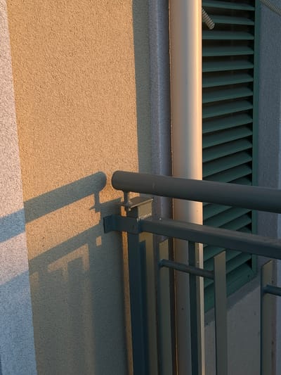 The image is a close-up shot of a textured exterior wall, likely part of a building in Chernomorets, Bulgaria, with a shadow cast across it. A metal railing with vertical supports is visible in the foreground, obscuring part of the wall and a beige pipe that runs vertically alongside a green louvered window or vent. The wall has a rough, sandy texture and is illuminated by warm, low-angle sunlight, creating a distinct shadow of the railing on its surface. The shadow exhibits sharp geometric shapes, suggesting the light source is positioned to the left of the frame. The overall scene conveys an outdoor architectural detail, possibly a balcony or exterior wall. There are no people or animals present in the image.
