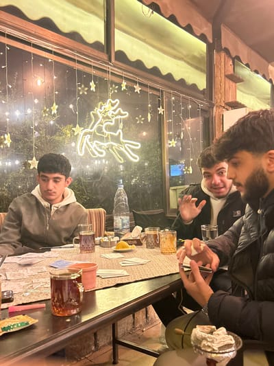 Three young men are seated at a table playing cards, with several glasses filled with liquid arranged around. A water bottle and ashtray are also visible. The setting appears to be indoors near a window adorned with star-shaped lights and a reindeer-shaped decoration. The men are dressed casually in jackets, with two facing the camera. The overall scene suggests a relaxed and friendly gathering.