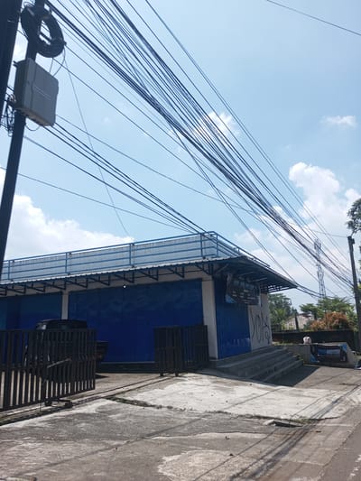 The image depicts an outdoor scene in Nawng Hkar, Myanmar. A low, single-story building with blue roll-up doors and a rooftop railing dominates the frame. A black vehicle is partially visible behind a metal gate in front of the building. Steps lead up to the building's entrance. A banner with the word "MOTOR" is placed on the side near the steps. Overhead, a dense network of power lines crisscrosses the sky. A utility pole with a junction box is on the left. The pavement is cracked and uneven. In the background, foliage and a cell tower are visible. The sky is partly cloudy.